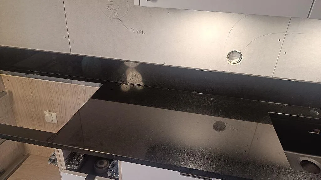 Lång arbetsyta i polerad Nero Assoluto granit med stänkkant, öppet skåp och diskho synlig under montering i Tibro.