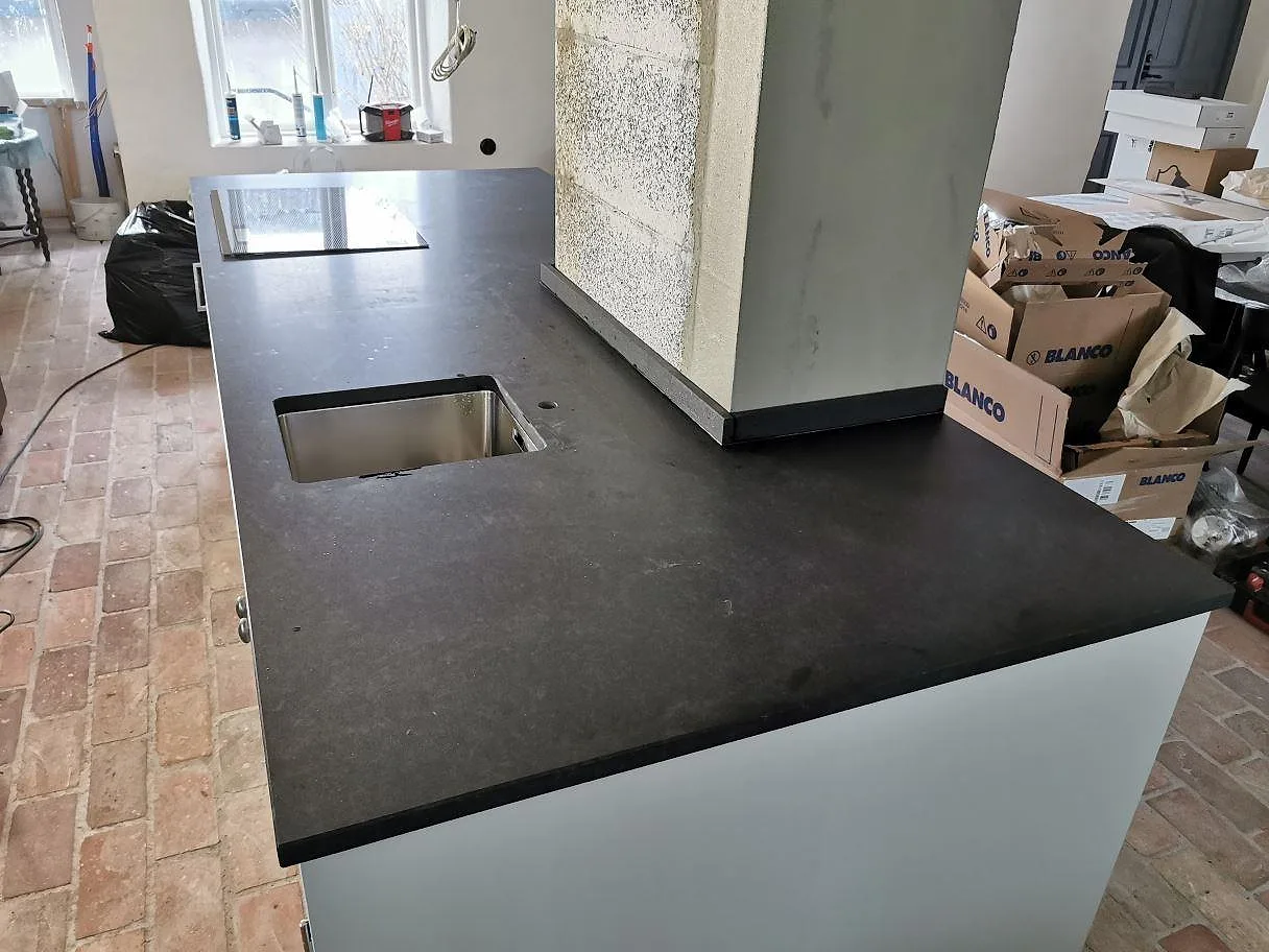 Köksbänkskiva i svart Nero Assoluto granit, matt yta, i ett L-format kök med underlimmad rostfri diskho, hällurtag och rött tegelgolv i Trelleborg.