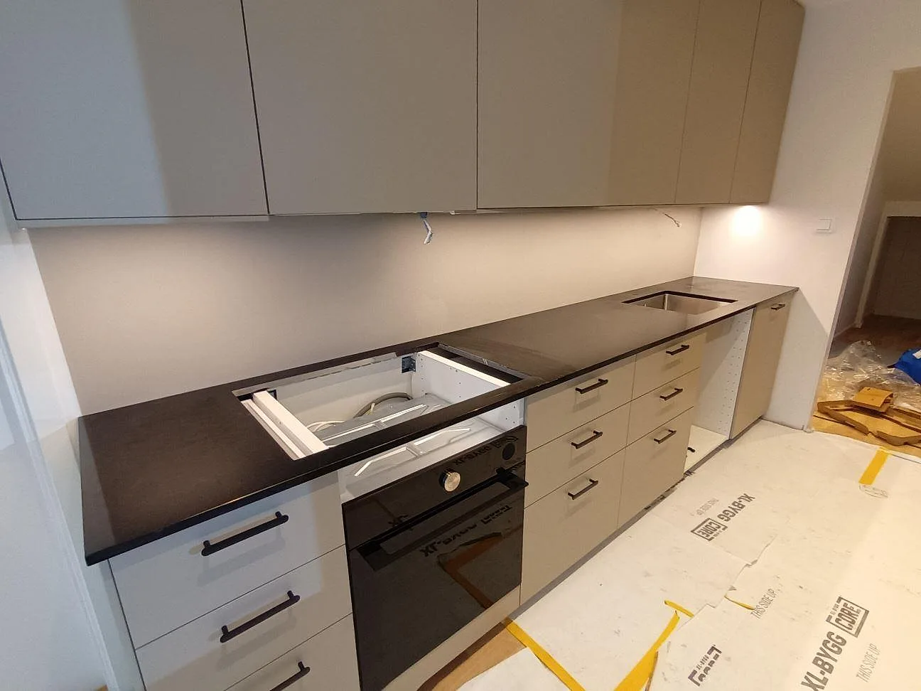 Köksbänkskiva i svart granit Nero Assoluto, polerad, längs en lång köksvägg med urtag för diskho och häll under montering i ett kök i Vällingby.