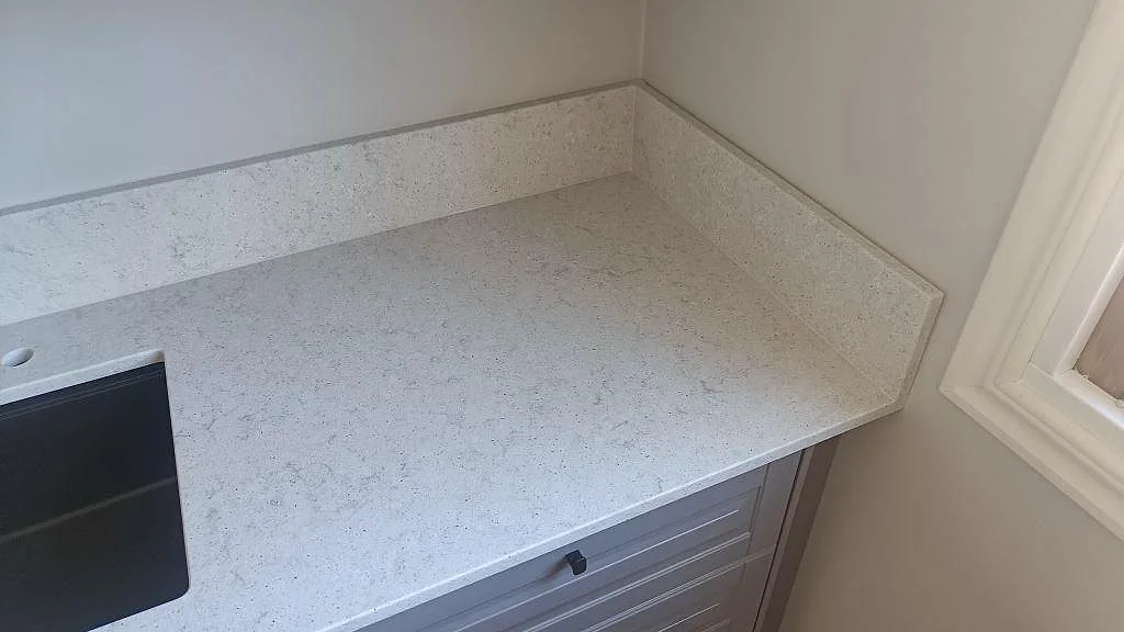 Köksbänkskiva i Quarella Bianco Savoia med uppvikt stänkkant och svart diskho i hörnet av ett kök i Nättraby.