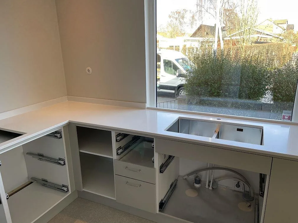 Köksbänkskiva i ljus Quarella Blanco Sal komposit, polerad, i L-form med underlimmad rostfri dubbelho under köksfönstret i ett kök i Limhamn.