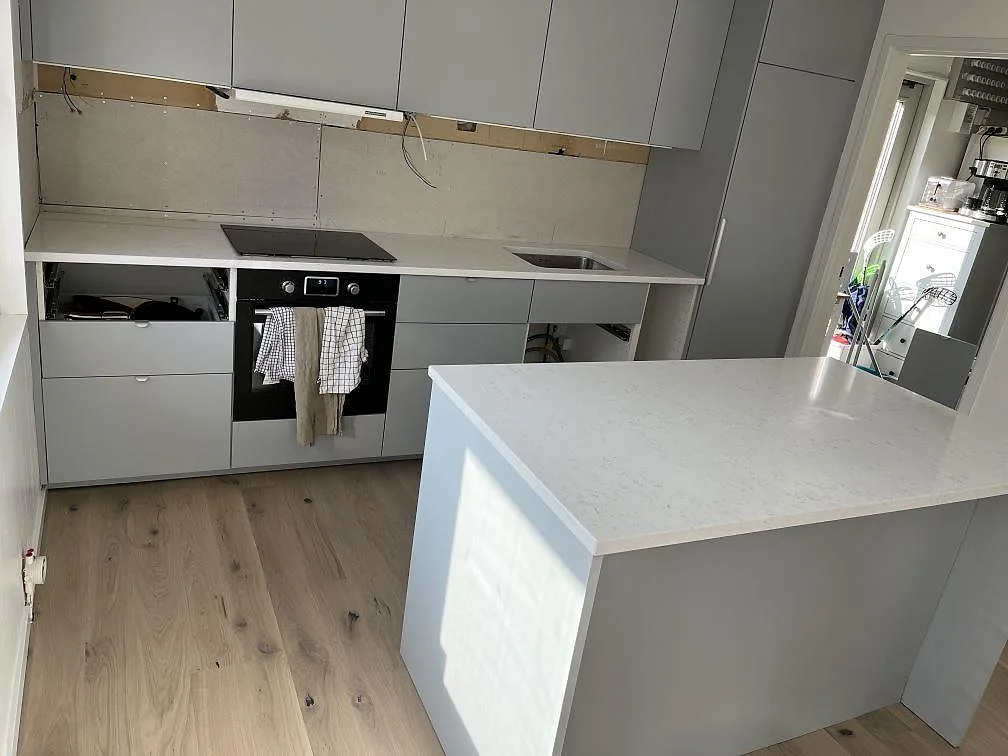 Köksbänkskiva i vit Quarella Navona kvartskomposit i ett kök i Lerberget under montering, med köksö i förgrunden och en bänk längs väggen med induktionshäll och underlimmad diskho.