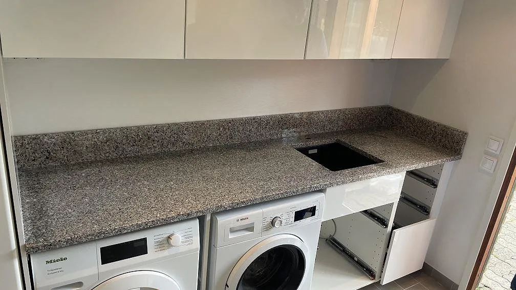 Tvättbänk i polerad Rosa Beta granit med svart drop-in-diskho, vit inredning och tvättmaskin i Hyllinge.