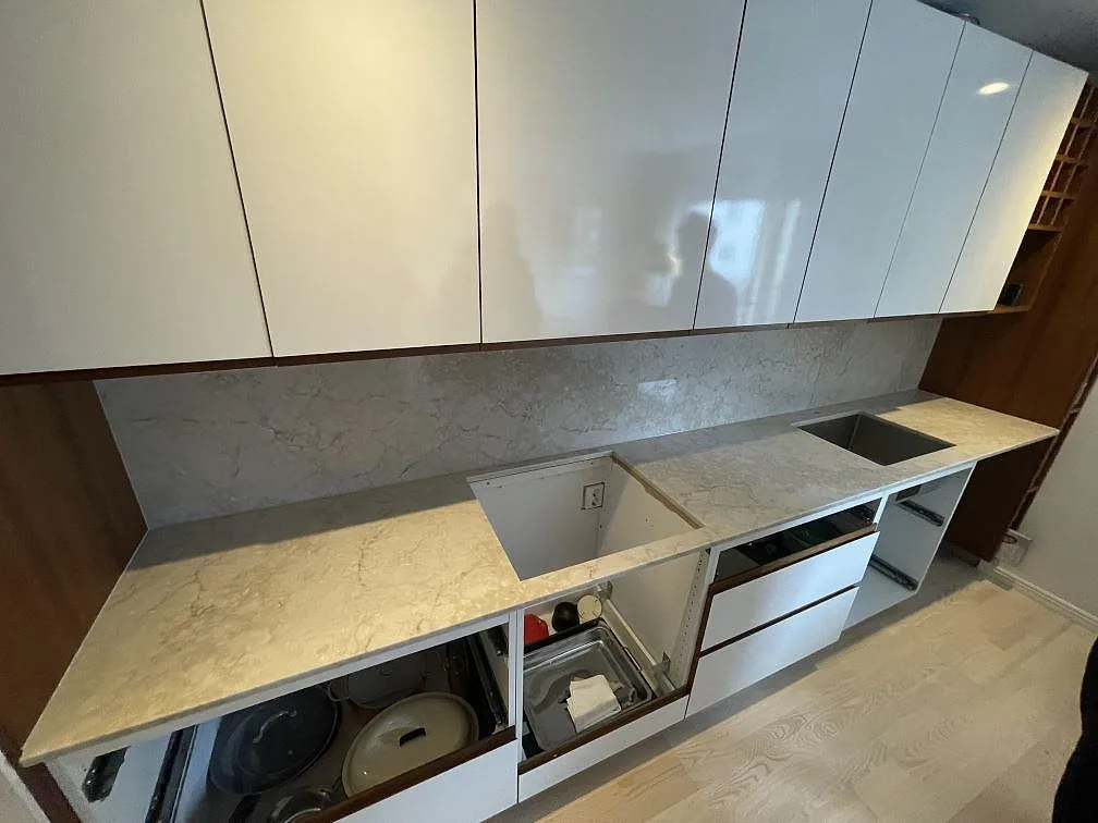 Köksbänkskiva i Royal Beige, polerad kvartskomposit i varm gräddbeige, längs en hel köksvägg med heltäckande grå stänkvägg, underlimmad diskho i rostfritt och urtag för häll, i ett kök under montering i Uppsala.