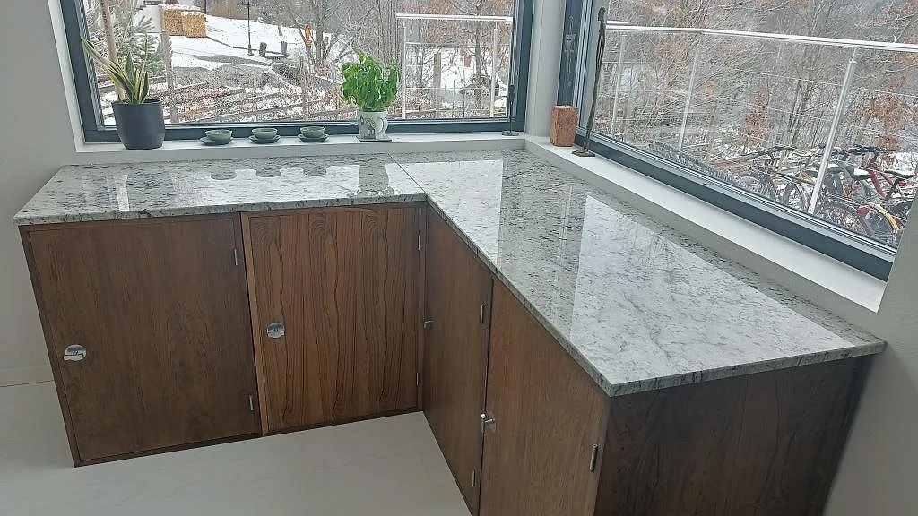 Köksbänkskiva i Antique White granit, polerad, L-formad och monterad på mörka träfronter med fönsterbänkar i ett kök i Sandared.