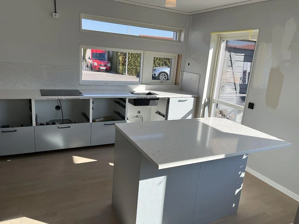 Köksbänkskiva och köksö i vit Silestone Bianco River, polerad, med infälld induktionshäll och uppvikt stänkskydd i samma sten i ett kök i Trelleborg.