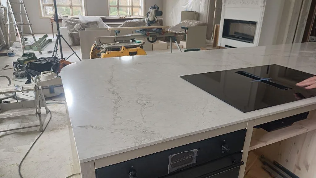 Bred vy över köksö med polerad Silestone Blanc Elysee som bänkskiva, svart häll och ljus ådring i ett kök i Allerum.