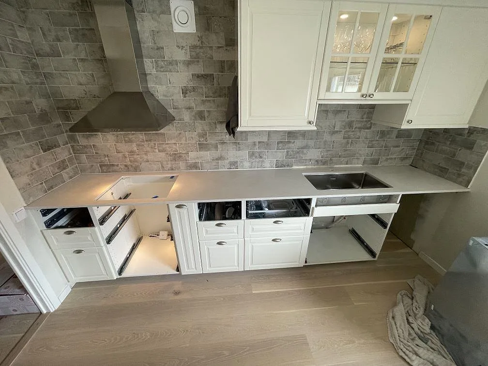 Köksbänkskiva i cremevit Silestone Blanco Maple 14, polerad 20mm, monterad under montering längs en L-formad köksbänk mot grått marmormönstrat tegelkakel, med vita ramluckor och rostfri spiskåpa i ett kök i Uppsala.