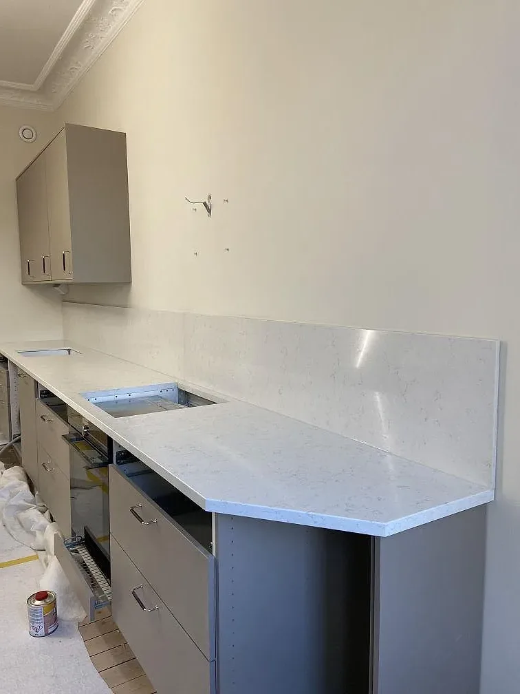 Köksbänkskiva i vit Silestone Blanco Orion med fint grått stänk, monterad bänkrad med vikt gavelparti och underlimmad diskho mot gröngrå stommar i ett kök under montering i Göteborg.