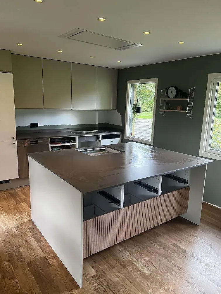 Köksbänkskiva i matt Silestone Brass Relish komposit med salviegröna skåp, träinredning och fönster i ett kök i Ängelholm.