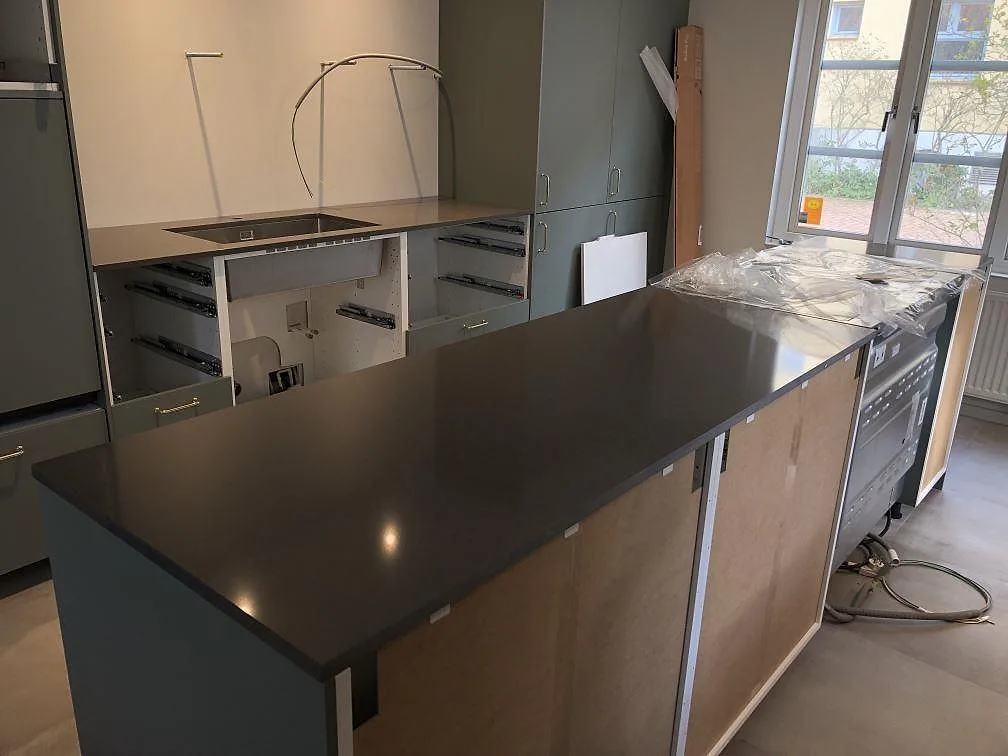 Köksbänkskiva och köksö i mörk brungrå Silestone Calypso, polerad, i ett kök under montering med urtag för häll och gröna stommar i Landskrona.