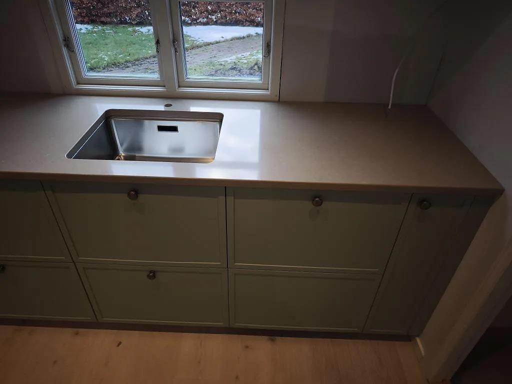 Köksbänkskiva i polerad Silestone Coral Clay med rostfri diskho vid fönster och gröna luckor i ett kök i Dalby.
