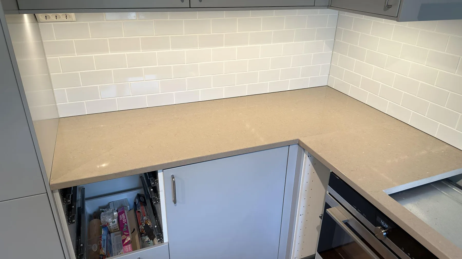 Hörnparti av polerad Silestone Coral Clay köksbänk med hälluttag och ljus kaklad vägg i ett kök i Ekerö.