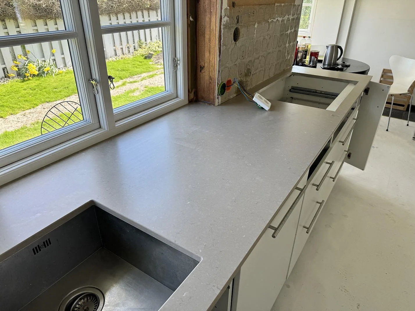 Lång köksbänkskiva i matt Silestone Coral Clay vid fönster, med rostfri dubbelho och hällurskärning i Falsterbo.