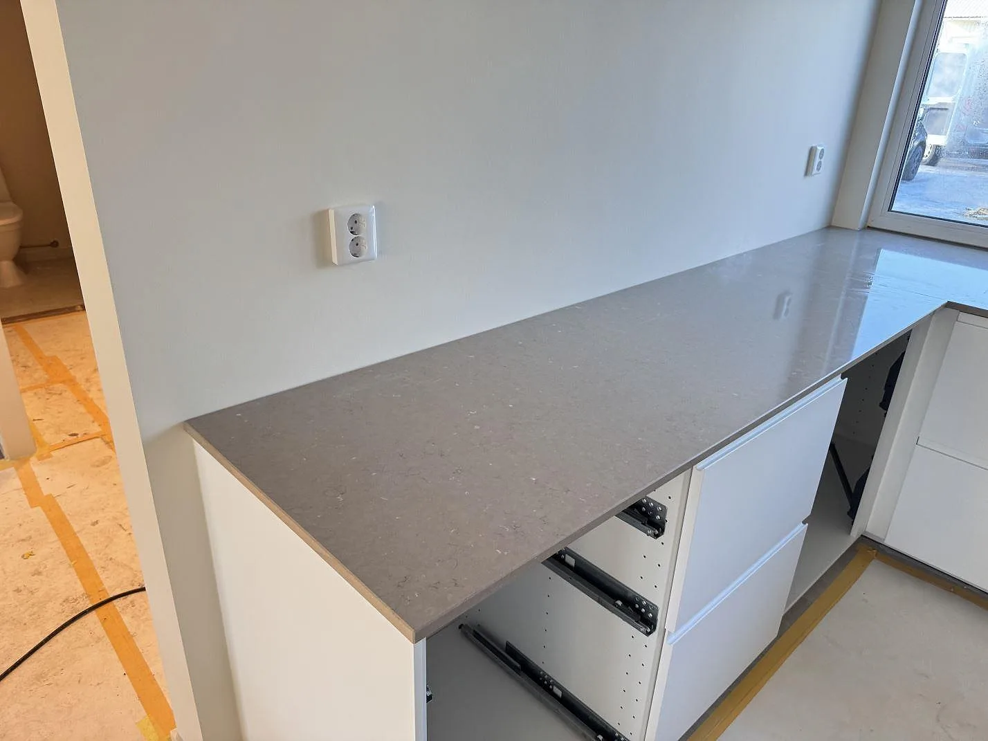 Köksbänkskiva i Silestone Coral Clay, polerad, längs en lång rak vägg i ett kök under montering i Halmstad, med öppna lådstommar och varm greige yta.