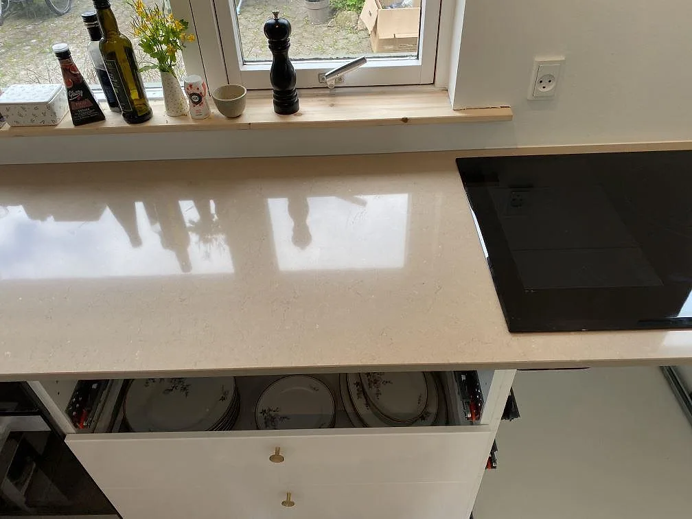 Köksbänk i Silestone Coral Clay, polerad gräbeige komposit, längs fönsterväggen med induktionshällens kant och öppna lådor i ett kök i Hørsholm.
