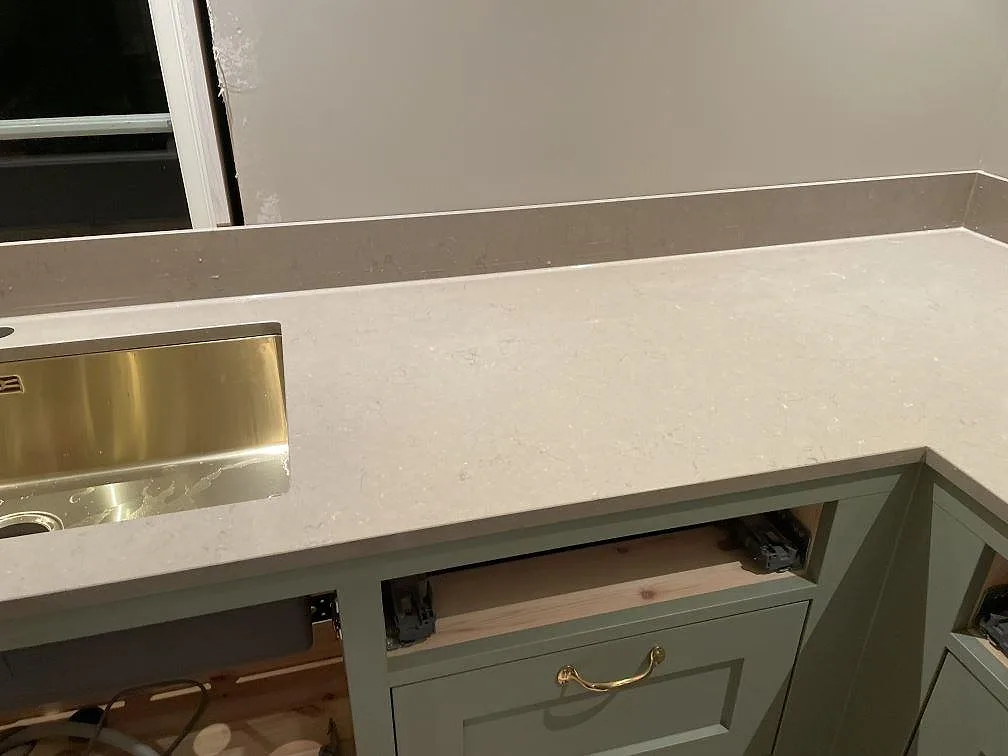 Köksbänkskiva i varm gråbeige Silestone Coral Clay med synlig ådring, stänkskydd och salviagröna stommar i Holsbybrunn.