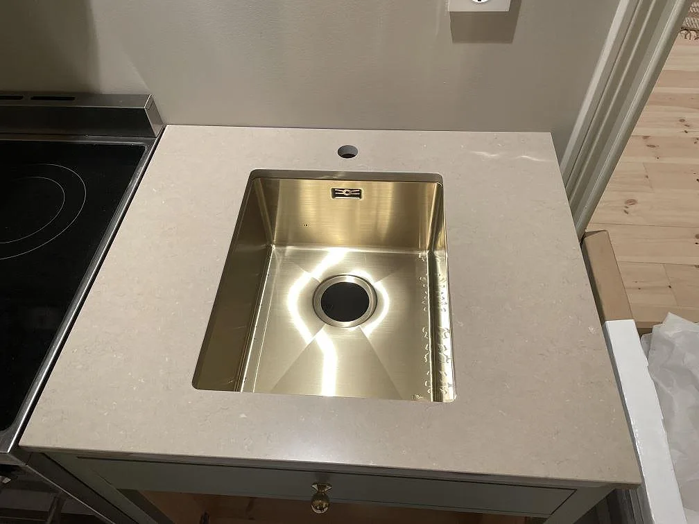 Detalj av underlimmad mässingsfärgad diskho i polerad Silestone Coral Clay bänkskiva med spis bredvid i Holsbybrunn.