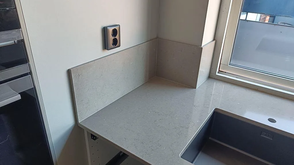 Närbild av polerad Silestone Coral Clay köksbänkskiva med gråbeige stänkskydd i samma material och mörk diskho vid fönster i Karlskrona.