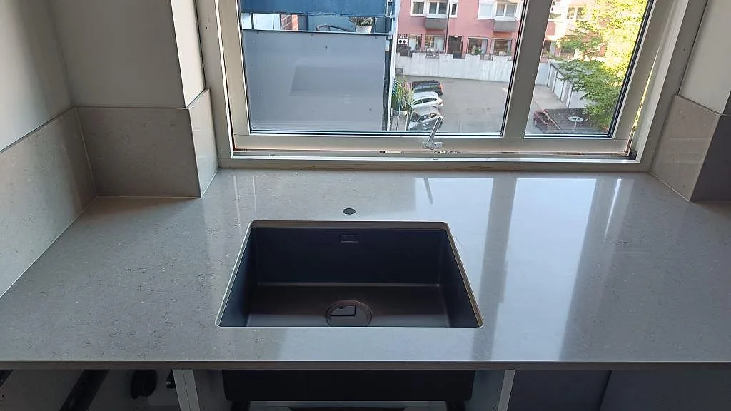 Frontvy av polerad Silestone Coral Clay bänkskiva med mörk diskho under stenen, kranhål och lågt stänkskydd vid fönster i Karlskrona.