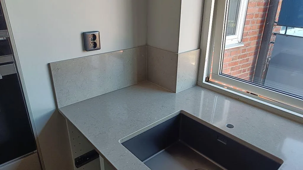 Polerad Silestone Coral Clay bänkskiva med uppvikt stänkskydd runt hörn, mörk diskho och fönsterljus i ett kök i Karlskrona.