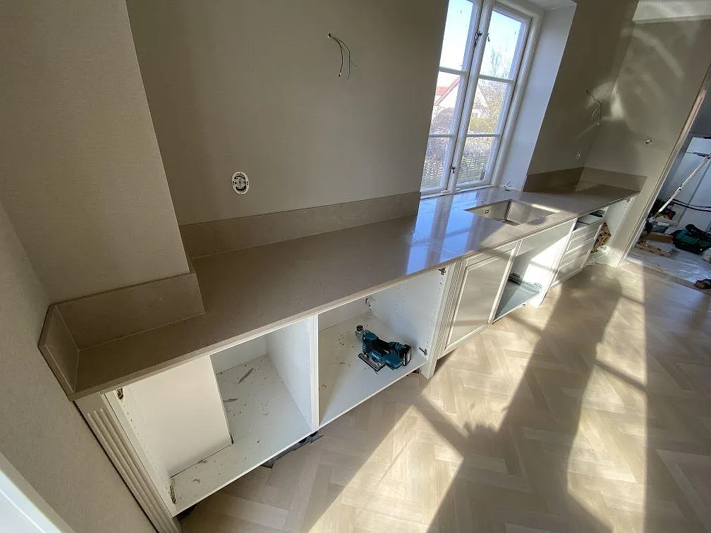 Lång väggbänk i polerad Silestone Coral Clay med diskho, fönster och lågt stänkskydd i ett kök under montering i Landskrona.