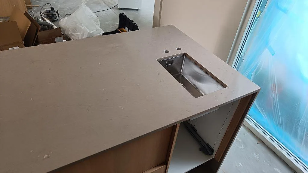 Närbild på polerad Silestone Coral Clay bänkskiva i kvartskomposit med synlig diskho, kranhål och ljusa stommar vid fönster i Lomma.