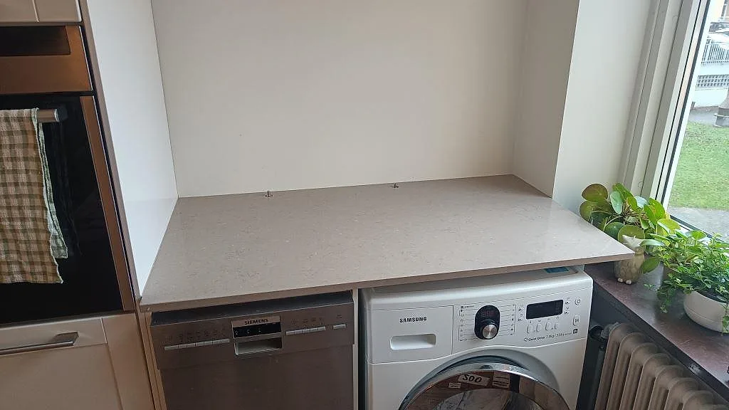 Rak polerad bänkskiva i Silestone Coral Clay, gråbeige kvartskomposit, över vitvaror vid fönster i Malmö.
