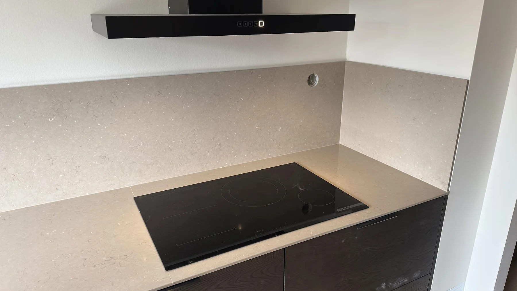 Polerad Silestone Coral Clay köksbänkskiva i Sollentuna med infälld induktionshäll och heltäckande stänkskydd i samma komposit under en svart köksfläkt.