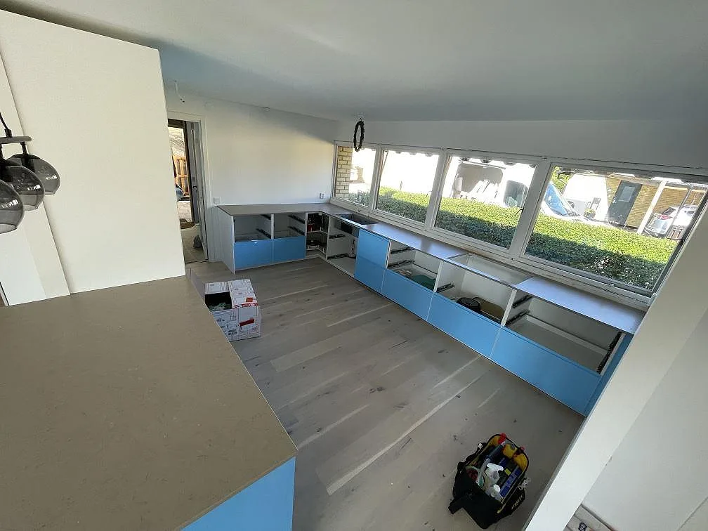 Översikt över kök i Staffanstorp med bänkskivor i matt Silestone Coral Clay längs stora trädgårdsfönster och blå stommar.
