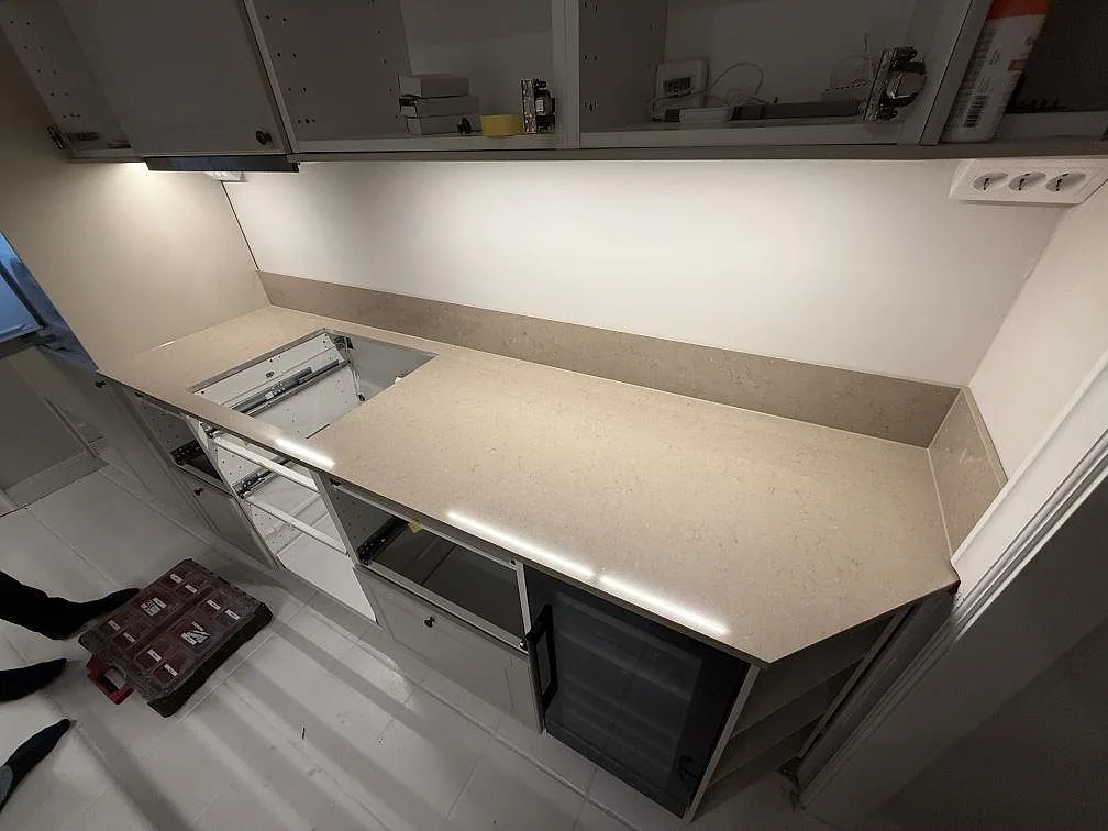 Lång köksbänkskiva i polerad Silestone Coral Clay med hällöppning, kort stänkskydd och öppna köksstommar under montering i Stockholm.