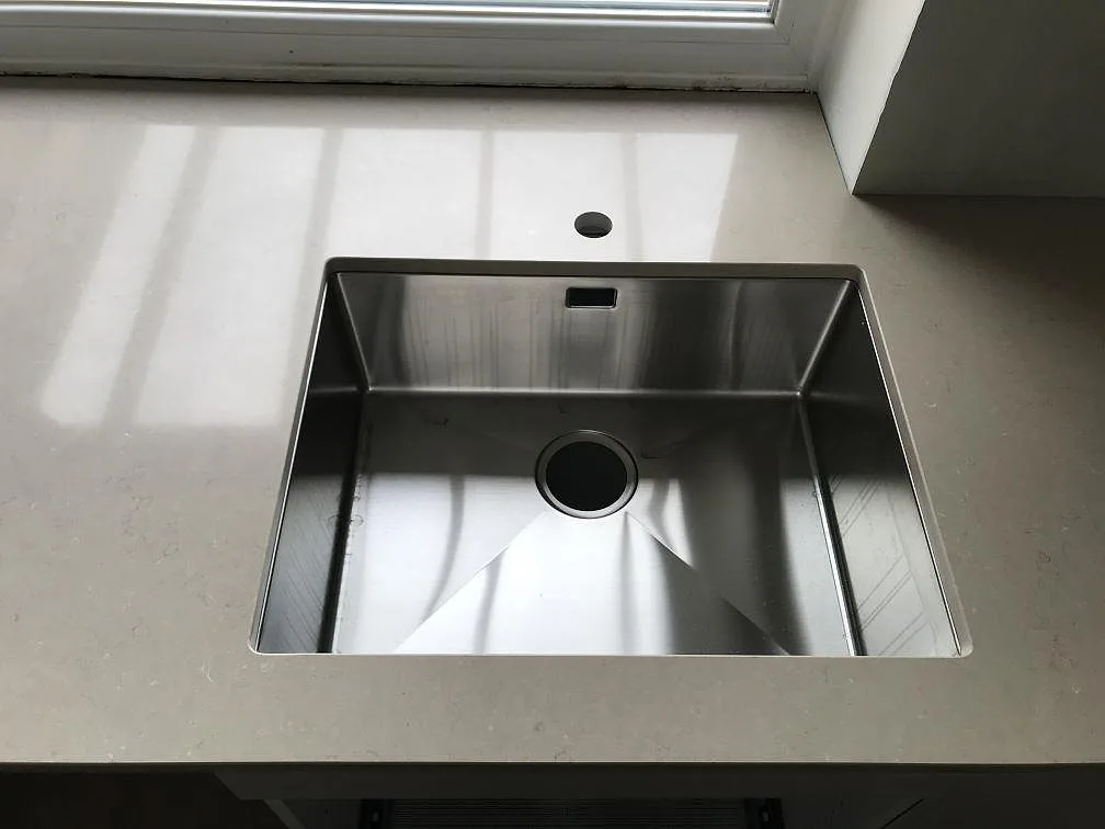 Underlimmad rostfri diskho infälld i polerad köksbänkskiva av Silestone Coral Clay under ett fönster i ett kök i Svalöv.