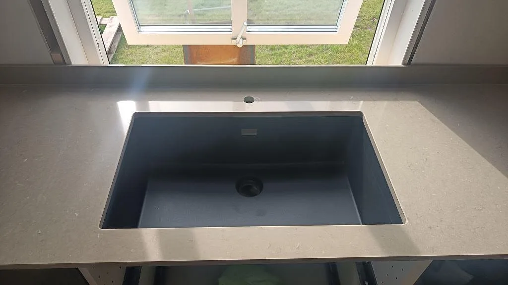 Närbild av svart diskho i polerad Silestone Coral Clay, gråbeige kompositbänkskiva med stänkskydd i väckelsång.