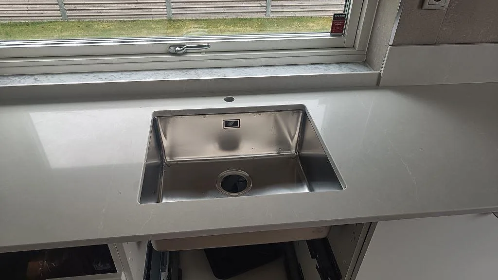 Rostfri diskho monterad i en polerad Silestone Desert Silver bänkskiva med fönsterljus och diskret ådring i Ängelholm.