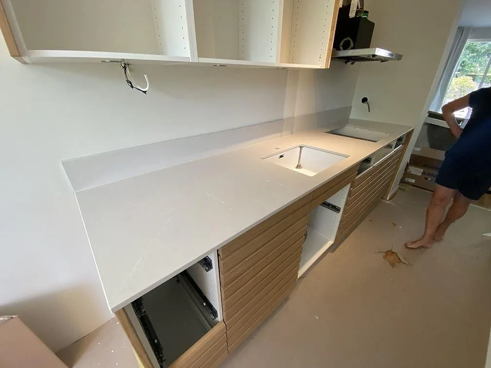 L-formad köksbänkskiva i polerad Silestone Desert Silver, 12 mm kvartskomposit, med låg stänkkant och träinredning i Halmstad.