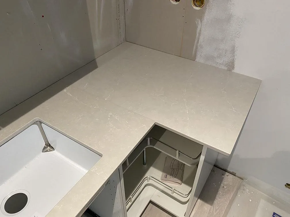 Hörndel av köksbänkskiva i Silestone Desert Silver, polerad komposit, vid vit diskho och öppna köksstommar i Höllviken.