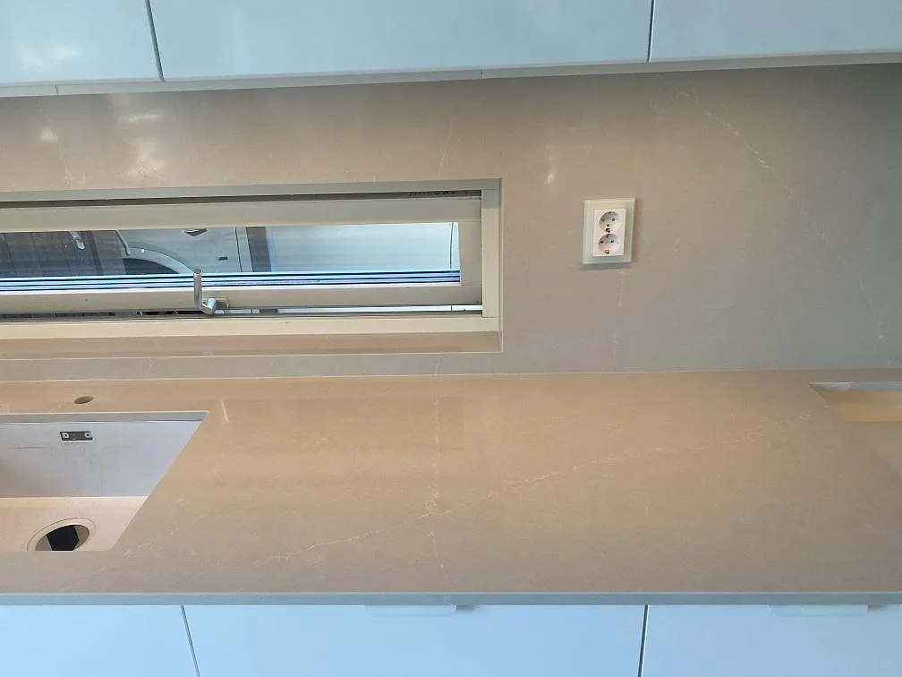 Bänkskiva i Silestone Desert Silver längs väggen med stänkskydd, urtag för diskho och häll i ett kök i Tygelsjö.