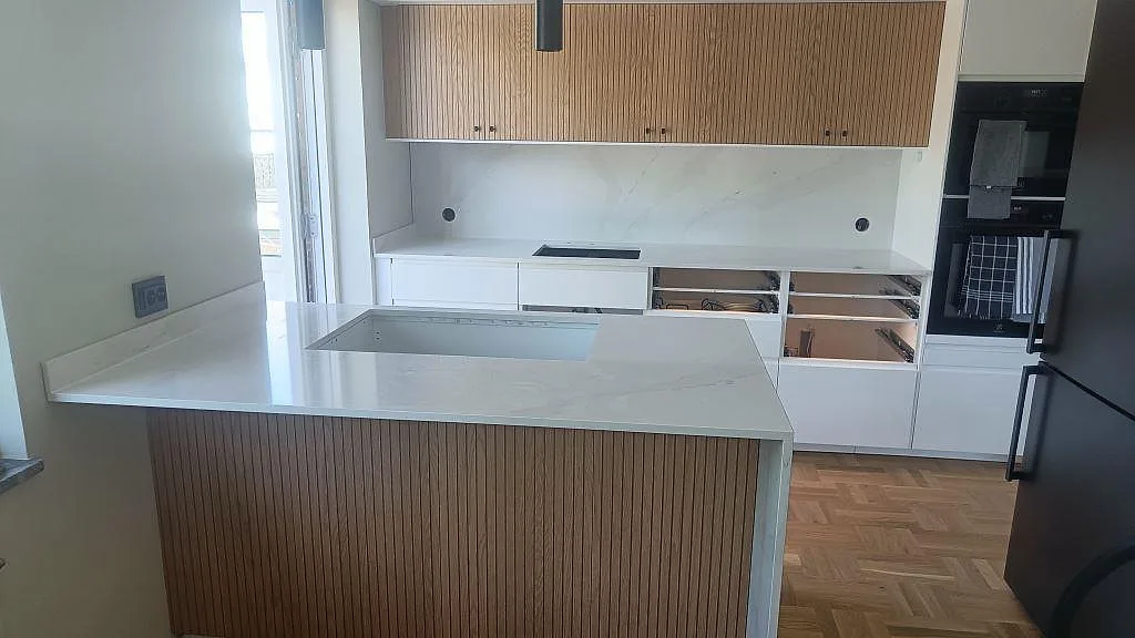 Polerad köksbänkskiva i vit Silestone Eternal Calacatta Gold med låg stänkkant, träspjälad halvö och köksinredning i Limhamn.