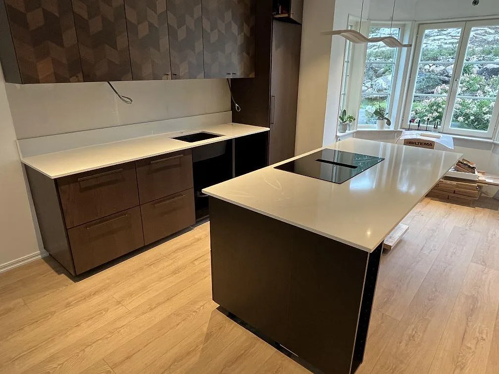 Köksbänkskivor i polerad Silestone Eternal Statuario med köksö, svart induktionshäll och mörkbrun träinredning i Rönninge.