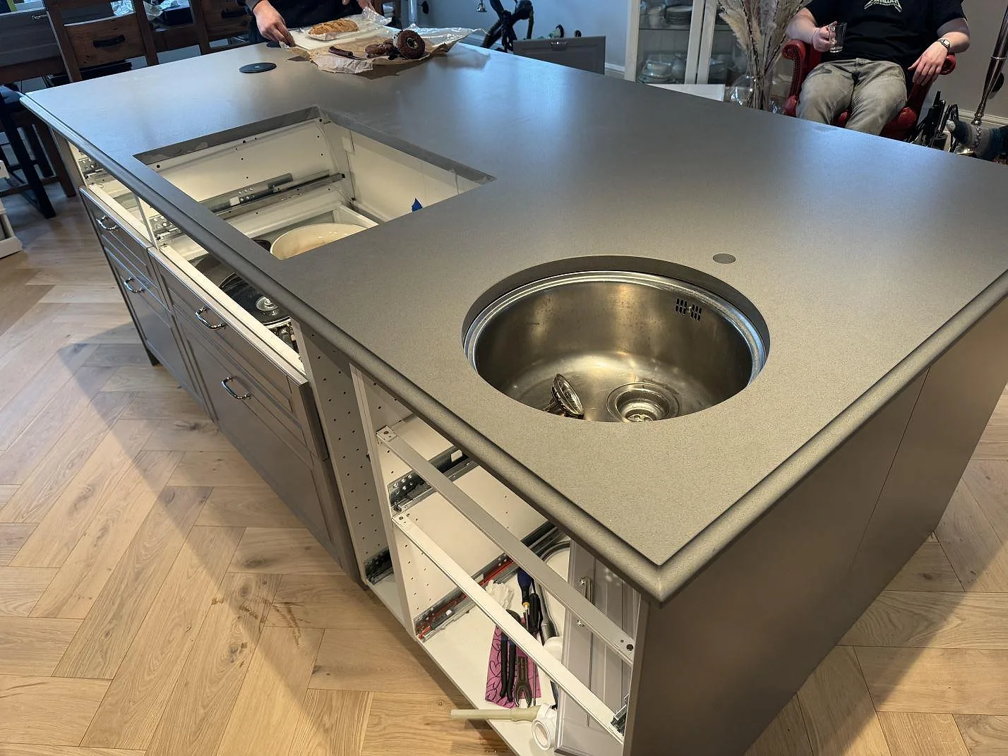 Köksö i Silestone Gris Expo, Mocka, med rund rostfri prep-ho, hälluttag och öppna stommar under montering i Sölvesborg.