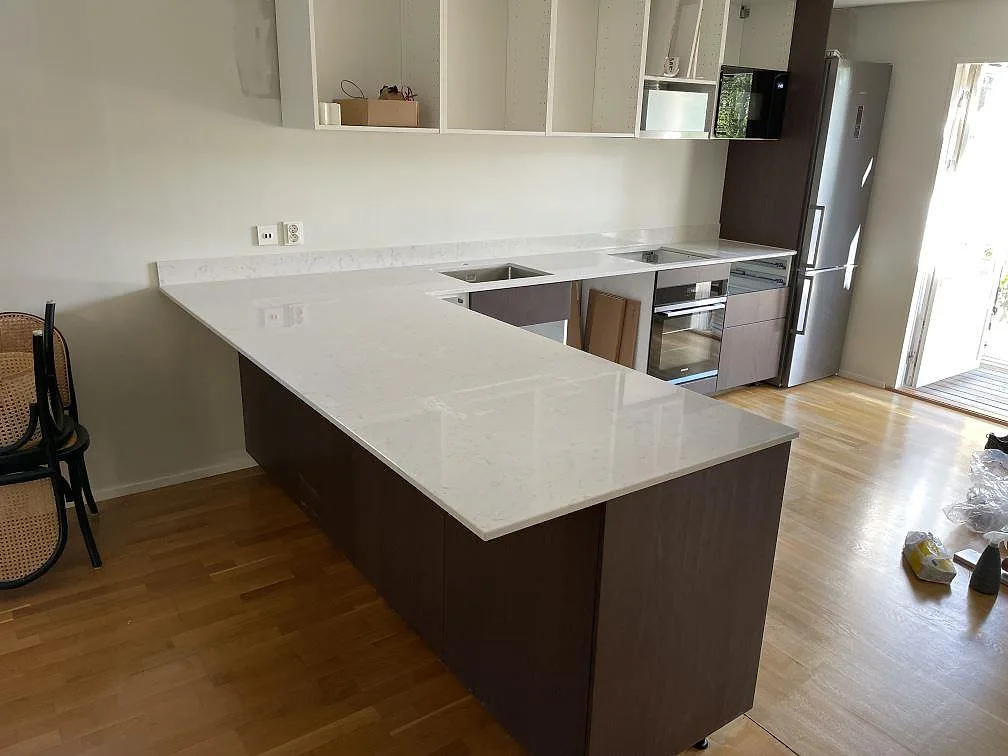 Köksbänkskiva i polerad Silestone Lagoon, vit kvartskomposit, på en halvö med låg stänkkant och synliga urtag i ett kök i Lidingö.