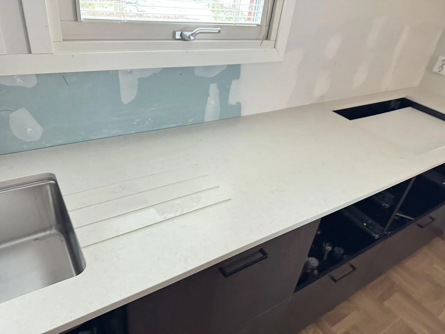 Köksbänkskiva i Silestone Linen Cream, matt cremevit komposit, längs en vägg under fönster med underlimmad diskho, avrinningsräfflor och urtag för häll i ett kök i Dalby under montering.