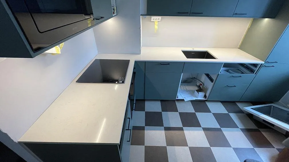 L-format kök i Malmö med polerad köksbänkskiva i Silestone Linen Cream, ljus kvartskomposit, samt svart häll och rostfri diskho under montering.