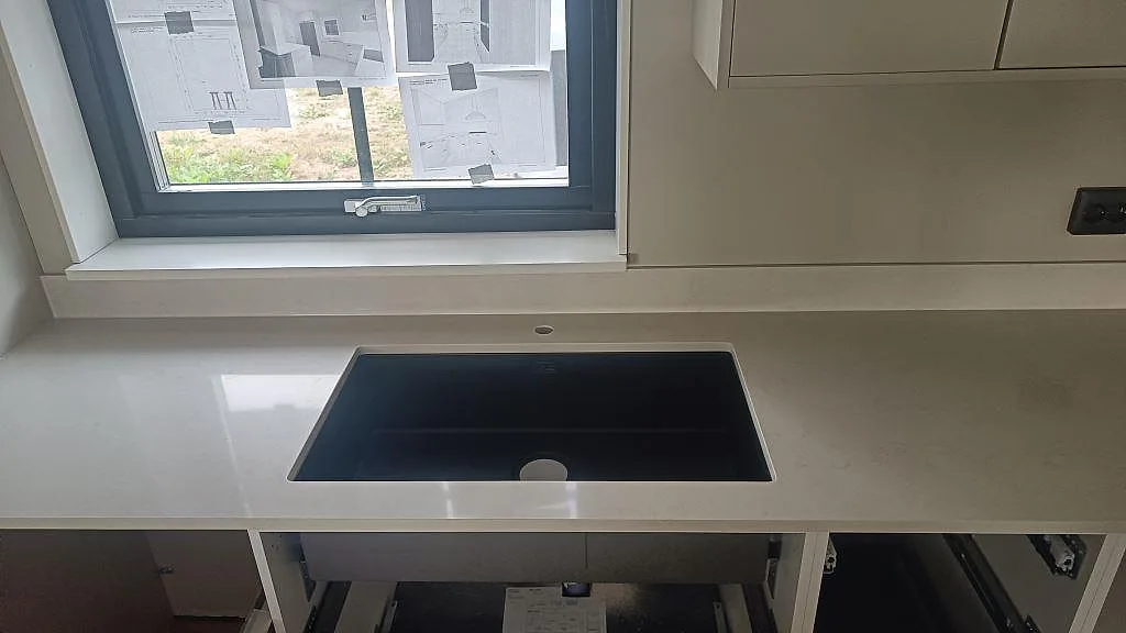 Rak köksbänkskiva i polerad Silestone Linen Cream med fönsterbänk, låg stänkkant och mörkt urtag i ett kök i Tågarp.