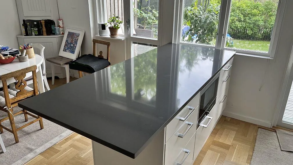 Polerad köksbänkskiva i mörkgrå Silestone Marengo kvartskomposit som halvö på vita skåp i ett kök med trägolv och trädgårdsfönster i Lidingö.