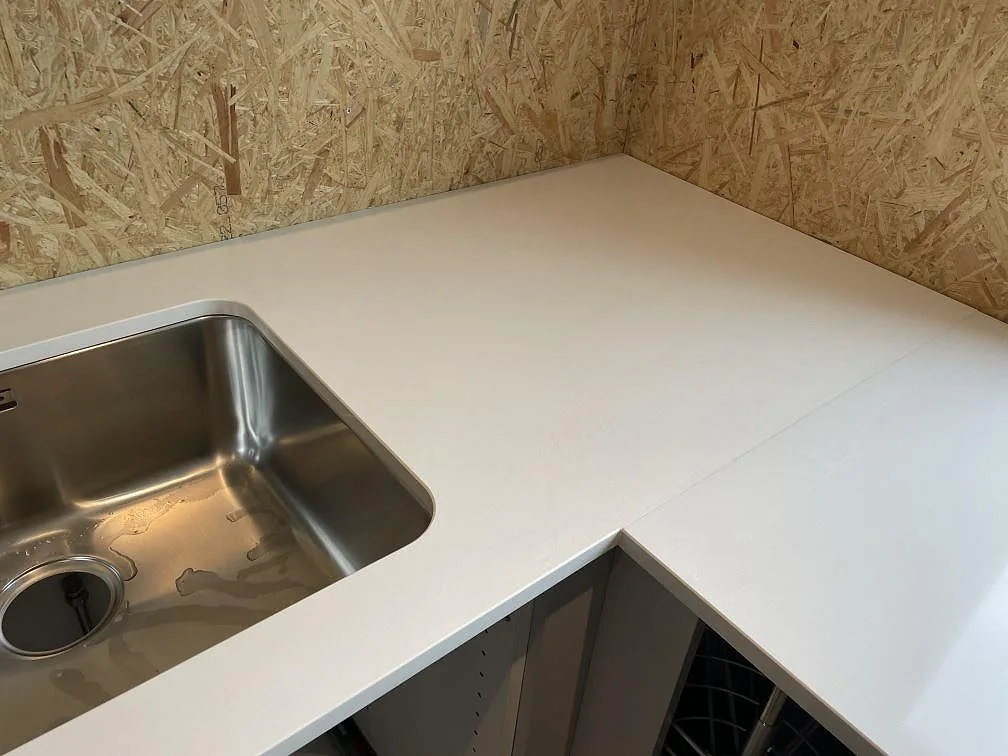 Hörndel av L-formad köksbänkskiva i polerad Silestone Miami White med underlimmad rostfri diskho och OSB-skiva bakom i Lyckeby.