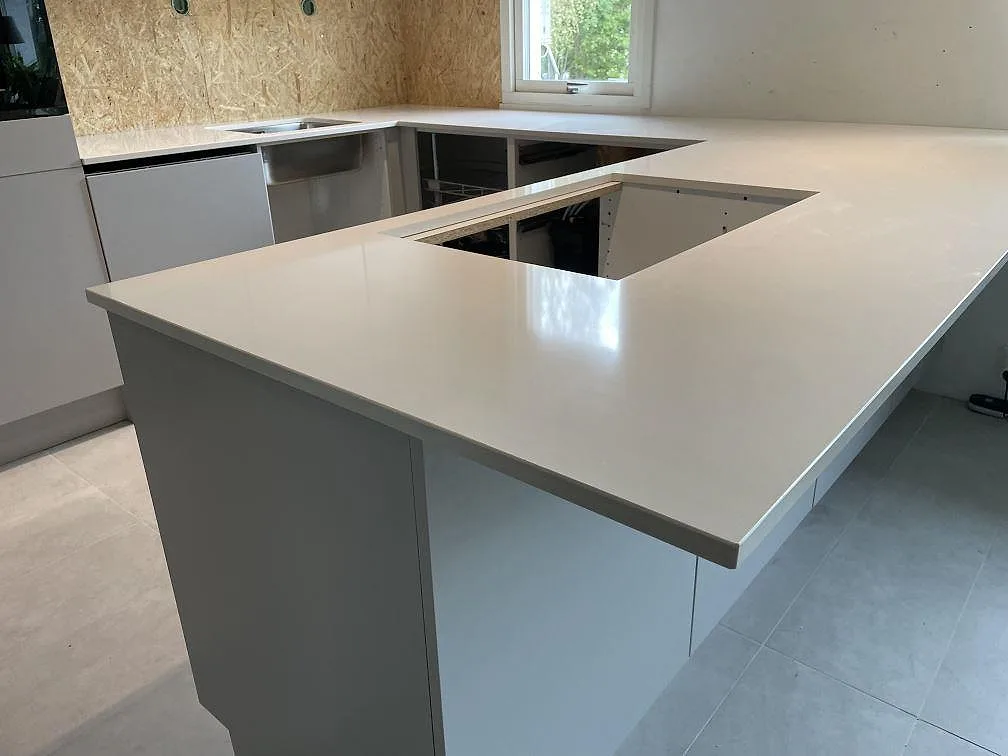 L-formad köksbänkskiva i polerad Silestone Miami White komposit med rektangulär urskärning, rostfri diskho och fönster i ett kök i Lyckeby.