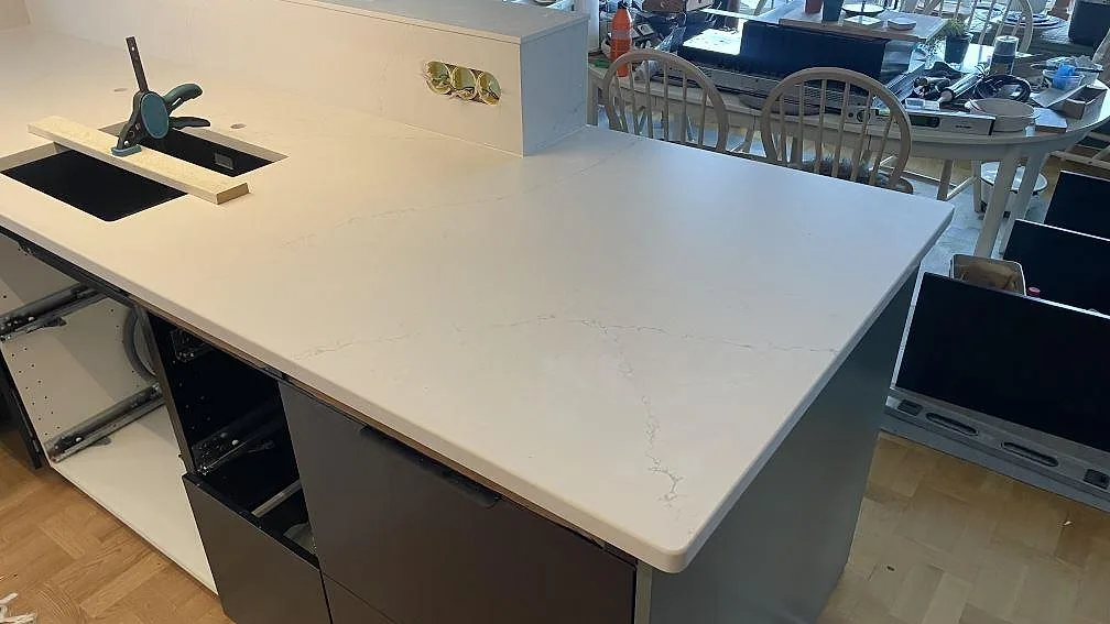 Stor arbetsyta på köksö i matt Silestone Siberian frost med rundat hörn, svart diskho under montering och stenpanel med uttag i Jönköping.