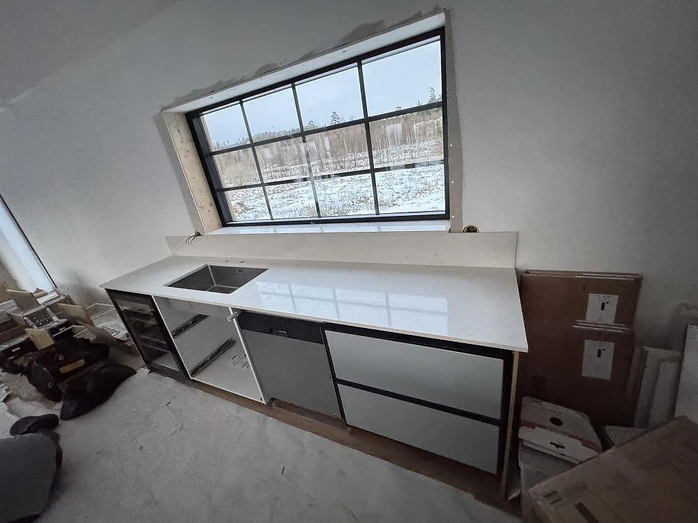 Bred vy över polerad köksbänkskiva i Silestone Snowy Ibiza med diskho och lång fönstervägg under montering i Alvesta.