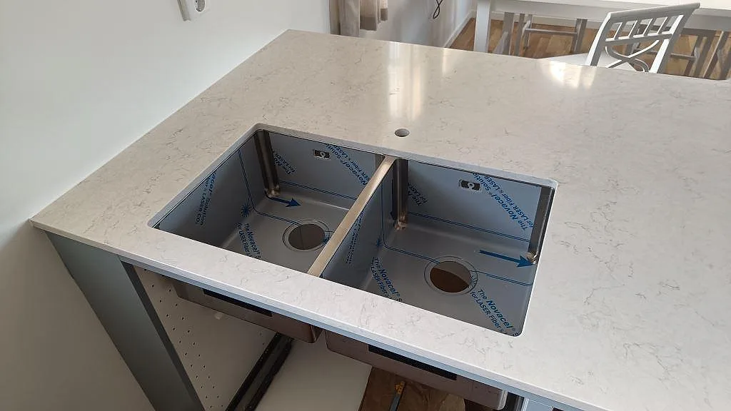 Närbild av köksö i polerad Silestone Snowy Ibiza, vit kvartskomposit med dubbel rostfri diskho under montering i Båstad.